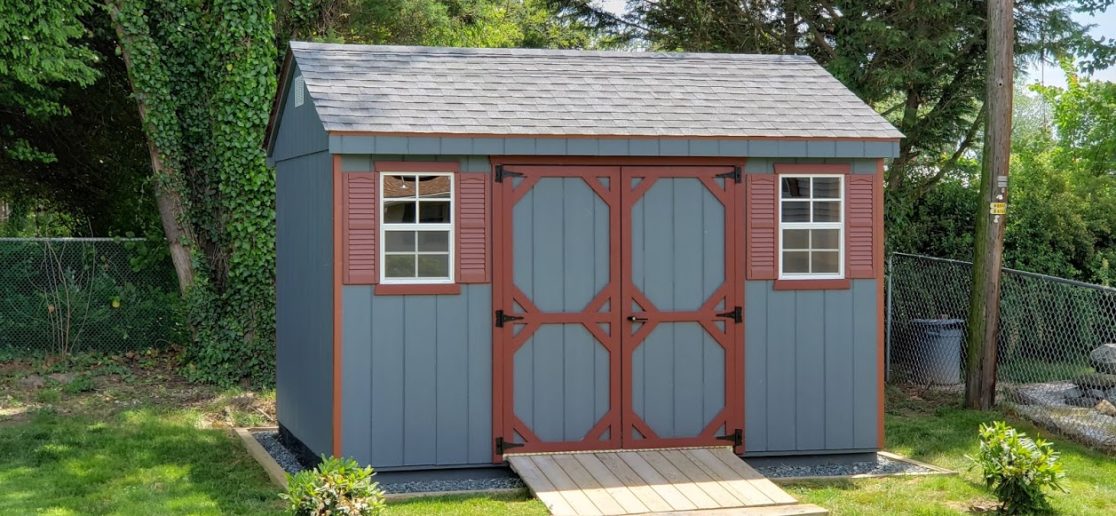 Blue Cottage Series Shed with dark red trim, double doors, 2 white windows with dark red shutters, a ramp, and light gray asphalt roofing.